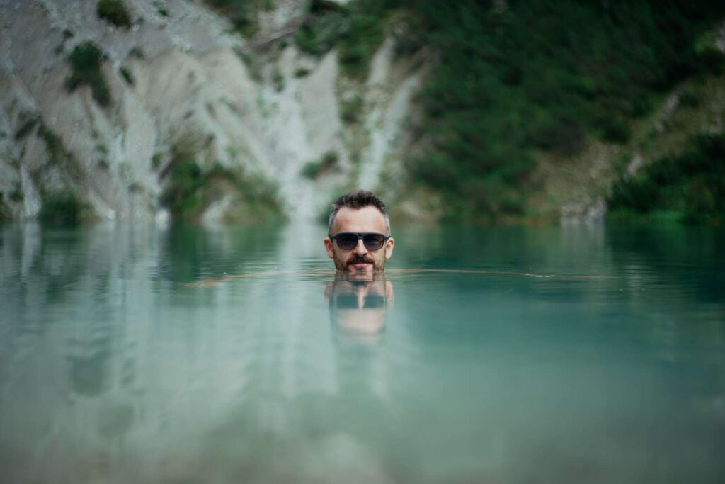 Mann-mittleren-Alters-mit-Sonnenbrille-schwimmt-in-klarem-Bergsee-vor-verschwommenem-Gebirgshintergrund