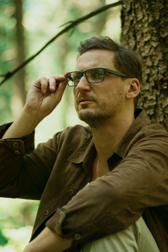 Man with green eyes adjusting glasses while sitting in a forest